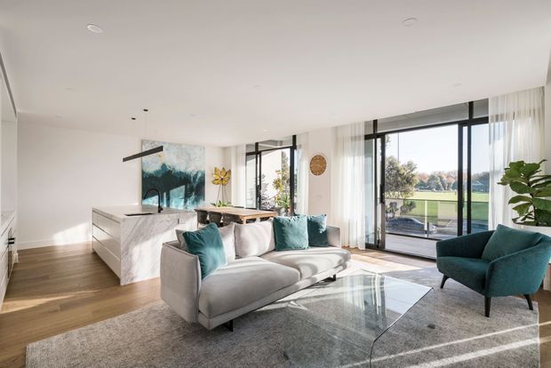 Bright open-plan living room with a contemporary kitchen visible behind it. The space features a large pale sectional sofa, teal accent cushions and armchair, a glass coffee table, warm timber flooring, light walls, floor-to-ceiling black-framed glazing, sheer curtains, and a marble-look kitchen island. The glazing overlooks a green open landscape, while a large abstract artwork and indoor planting add decorative accents.