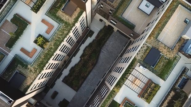 Aerial oblique view of a dense contemporary multi-storey residential development, showing flat-roofed buildings arranged around a central paved courtyard or access space. The roofs feature extensive planted green roofs and landscaped terraces, with light rendered facades, dark cladding elements, balconies, skylights, and rooftop seating areas.