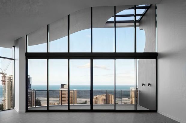 A bright, double-height contemporary interior with expansive black-framed floor-to-ceiling glazing overlooking a coastal urban skyline and ocean. The room has pale smooth walls, a light timber-toned floor, a dramatic curved ceiling edge, and a roof skylight. The space is largely unfurnished, emphasizing the architectural volume, panoramic outlook, and abundant natural light.