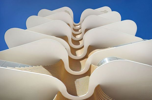 Upward-looking architectural view of a tall contemporary building with dramatic undulating balconies and layered, wave-like floor plates. The predominantly white facade contrasts strongly against a vivid blue sky, with warm-toned horizontal soffit and balcony detailing visible in the recesses.