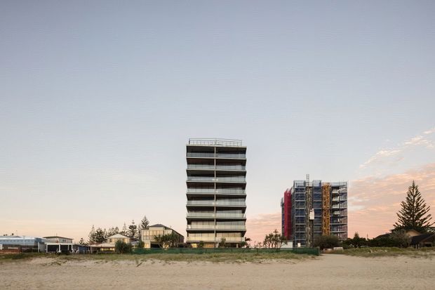 Wide exterior view of a beachfront or coastal urban development, showing two contemporary multi-storey apartment buildings beyond a sandy foreground. The taller central building has stacked horizontal balconies and dark glazing, while the building behind it features varied coloured vertical facade elements. Low-rise buildings, coastal vegetation, and a calm sky complete the scene.