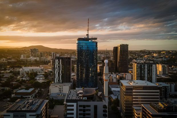 Aerial view of a dense urban skyline at sunset, featuring a prominent high-rise tower under construction with a crane, surrounded by modern apartment and commercial buildings. Low hills and a dramatic cloud-filled sky form the background.