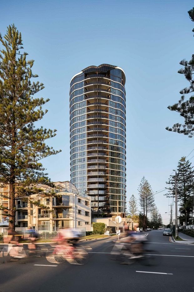 Street-level exterior view of a tall, modern residential apartment tower with a rounded cylindrical form, extensive curved glazing, continuous wraparound balconies, and a sculptural curved roofline. Lower-rise residential buildings, coastal-style pine trees, a paved urban street, and motion-blurred cyclists frame the tower.