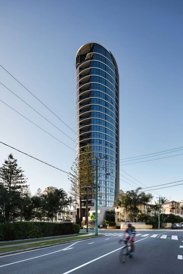 Street-level exterior photograph of a tall, slender contemporary apartment building with a rounded vertical form, extensive blue-tinted glazing, curved balconies, and dark structural framing. The tower stands on a landscaped urban/suburban street with marked road crossings and overhead utility wires.