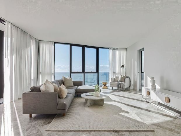 Bright, spacious contemporary living room with floor-to-ceiling glazing, sheer white curtains, a large grey sectional sofa, pale textured rug, round coffee table, sculptural decor, and a low white media console. The room overlooks a distant urban or waterfront view and receives abundant natural light.