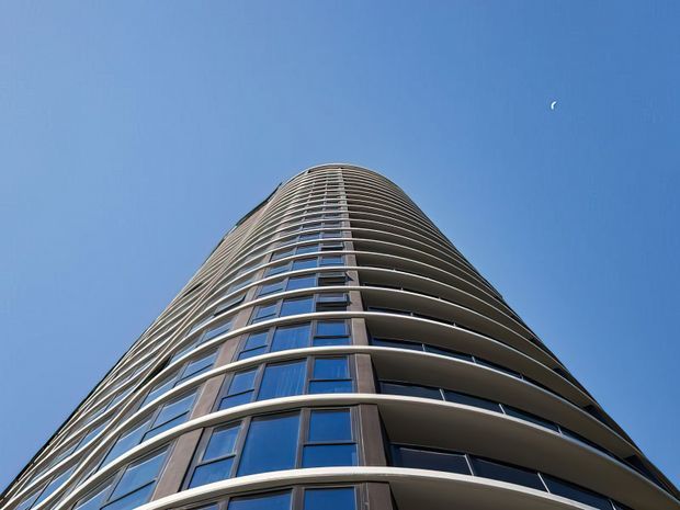Upward-looking exterior view of a tall curved residential or commercial tower against a clear blue sky. The facade features repeated stacked balconies with smooth rounded edges, dark glazed window panels, and vertical dark structural or cladding elements, creating a sleek contemporary high-rise form.