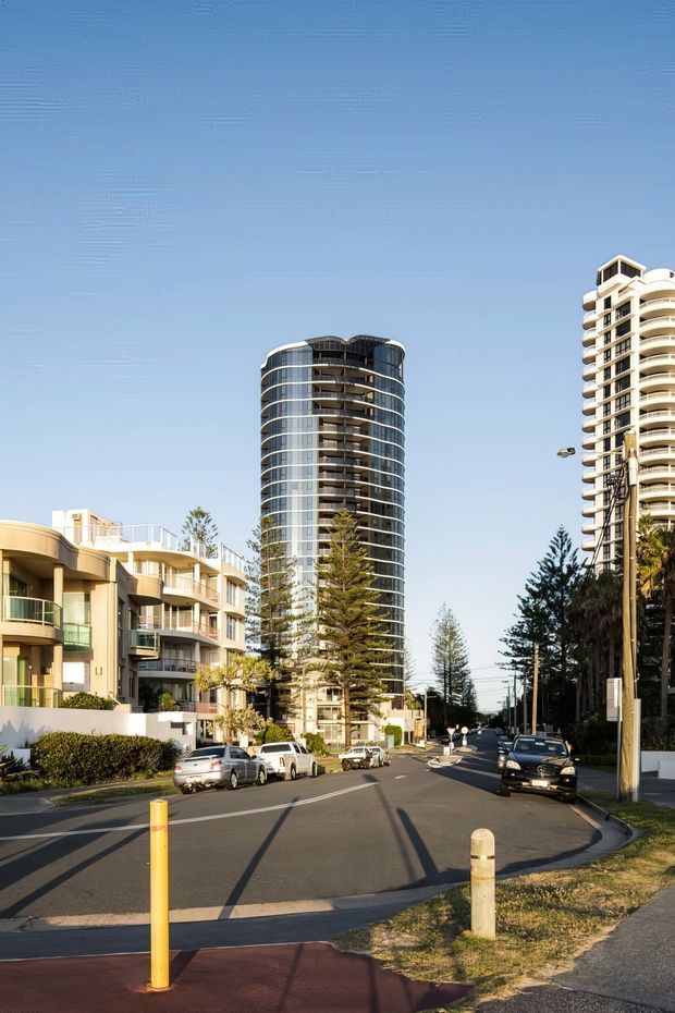 Street-level view of a sunny urban residential area with several tall apartment towers, rounded balconies, extensive glazing, and lower-rise apartment buildings lining a paved road. Mature trees, parked cars, street lighting, and pedestrian infrastructure are visible under a clear blue sky.