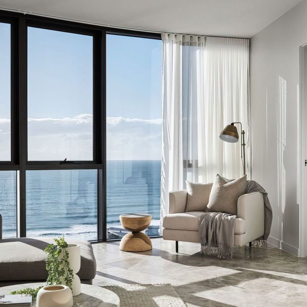 Bright contemporary living room with floor-to-ceiling black-framed glazing overlooking the ocean. The room features a cream upholstered armchair, soft neutral textiles, a sculptural wooden side table, sheer white curtains, pale stone-look flooring, and abundant natural light.