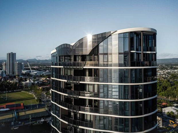 Aerial view of a contemporary multi-storey residential tower with a rounded, sculptural form, extensive floor-to-ceiling glazing, dark facade panels, curved balconies, and horizontal white balcony bands. An urban cityscape and sports fields are visible in the background.