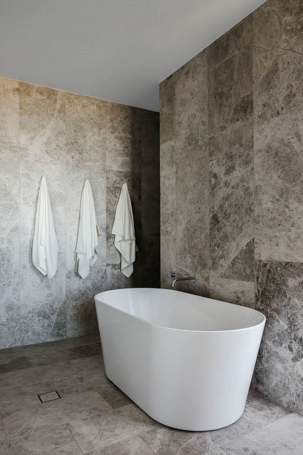 A refined contemporary bathroom featuring a freestanding white oval bathtub, large-format grey and taupe marble-look stone tiles covering the walls and floor, a wall-mounted tap, and three white towels hanging on the tiled wall. The space has a clean, minimalist composition with a neutral, slightly moody palette.