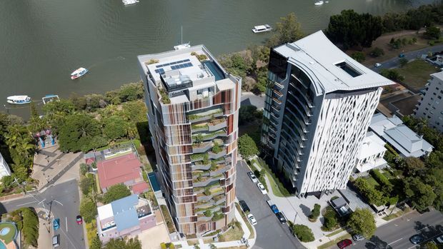 Aerial view of a modern high-density waterfront development, showing two contemporary multi-storey apartment buildings beside a river or harbour. The buildings feature flat roofs, extensive balconies, glazed facades, and distinctive vertical facade screens. Surrounding landscaped areas, roads, boats, and recreational courts are visible.
