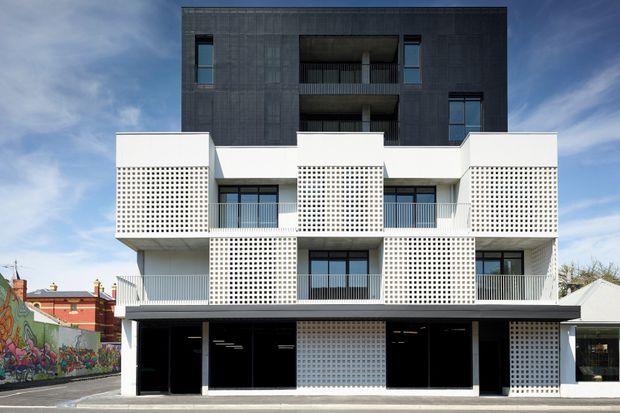Front elevation of a contemporary multi-storey urban apartment or mixed-use building. The lower levels feature white perforated masonry screens, projecting balconies, dark glazing, and a broad black canopy, while the upper storey is clad in dark grey panels with recessed balconies. A colourful mural and neighbouring low-rise buildings appear at the edges of the streetscape.