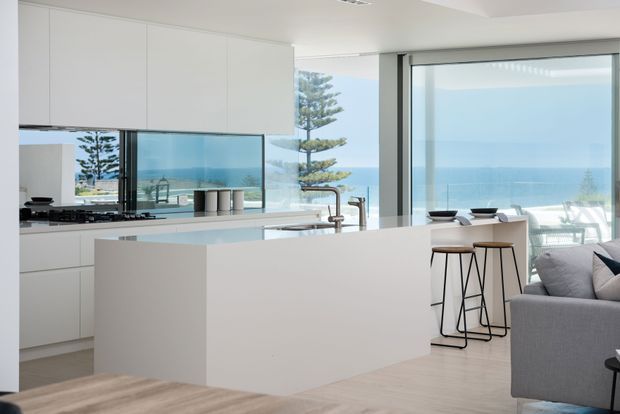 Bright open-plan coastal interior featuring a minimalist white kitchen with a large island, integrated cabinetry, reflective splashback, and a timber-toned floor. Floor-to-ceiling glazing provides expansive ocean views, while a small bar seating area and part of a grey sofa create a connection to the living space.