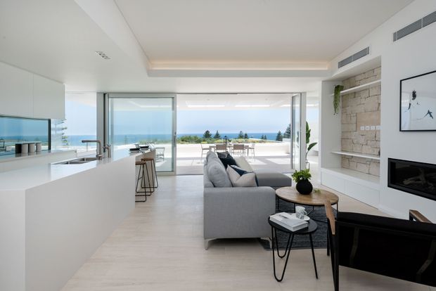 Bright open-plan contemporary living space with a white minimalist kitchen and island in the foreground, opening directly to a lounge and expansive ocean-view terrace. The room features floor-to-ceiling glazing, pale flooring, a grey sofa, black-framed furniture, a built-in fireplace, and a textured light-stone feature wall. The overall atmosphere is airy, refined, and coastal.