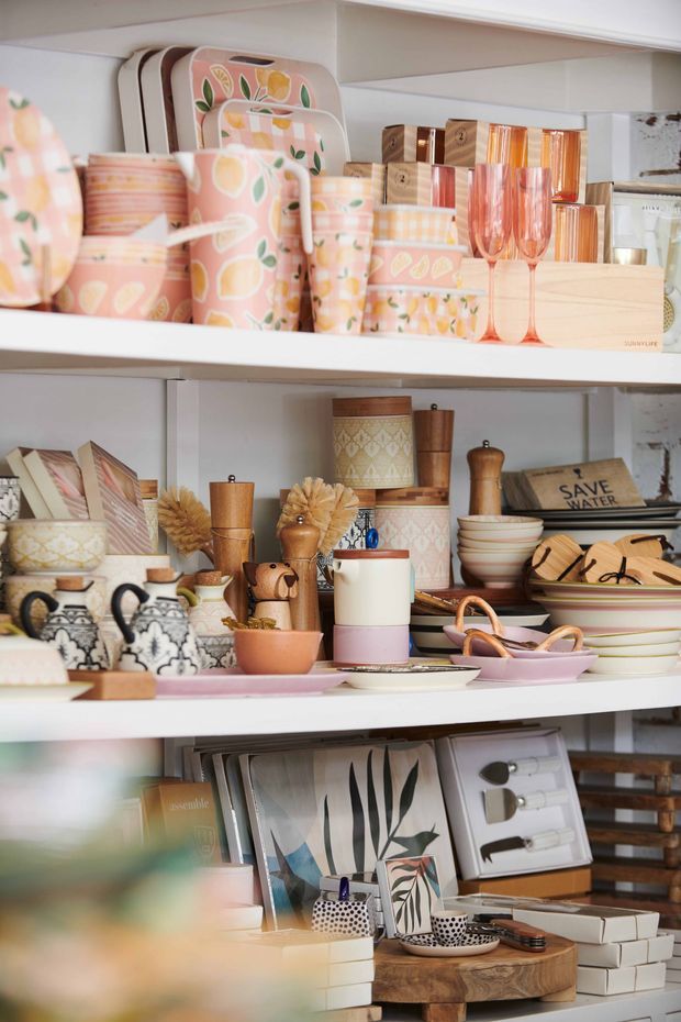 A close-up view of styled retail shelving displaying colourful kitchenware, tableware, serving trays, glassware, ceramics, wooden utensils, framed prints, books and decorative accessories. The shelves are white, with a warm pastel palette of peach, pink, cream, terracotta and natural wood. The image resembles a curated homewares shop display or showroom.