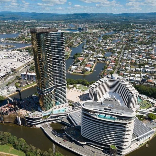 Aerial view of a dense urban waterfront precinct featuring two prominent high-rise buildings with curving balconies and extensive glass façades, surrounded by canals, residential development, roads, landscaped areas and water features. The image is dominated by the exterior architecture and broader site context.