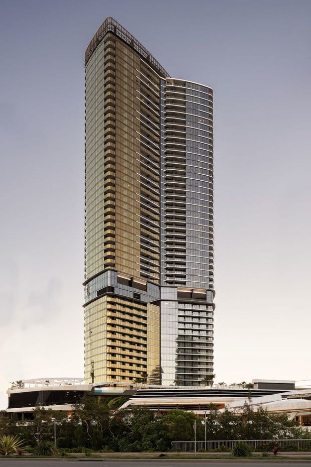 Photograph of a tall contemporary high-rise tower with extensive glazed curtain walls, curved balcony edges, and a mixed-use podium at its base. The building has a dark bronze and grey facade, strong vertical proportions, and a prominent rooftop structural screen, set in an urban landscaped setting.