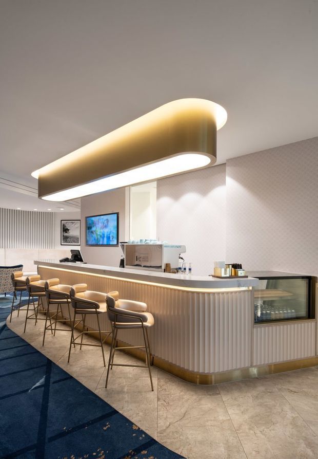 A refined commercial hospitality counter or café reception area with a long fluted-front service desk, integrated display case, upholstered bar stools, patterned wall covering, and a large sculptural illuminated pendant. The space combines warm metallic accents, pale stone-look flooring, and deep blue carpet in a polished contemporary fit-out.
