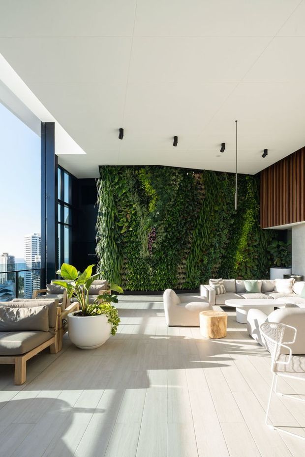 A bright, spacious contemporary living room with floor-to-ceiling corner glazing, pale timber-look flooring, white upholstered sofas and lounge chairs, and a large vertical living wall forming the main feature backdrop. Dark structural window framing, a timber slatted wall section, minimalist black track and pendant lighting, and a city skyline visible through the glazing complete the interior.