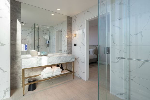 A bright, refined bathroom with full-height white marble-look wall surfaces, a large mirrored vanity wall, a floating stone benchtop on an open brass-toned metal frame, and neatly arranged towels beneath. A glass-enclosed shower occupies the right side, with a bedroom visible through the open doorway beyond. Recessed ceiling lights and a small wall sconce provide even illumination.