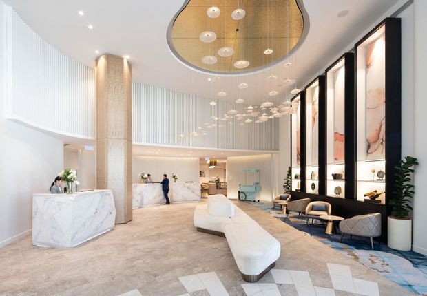 A spacious, high-end hotel or commercial lobby with a sculptural white reception desk, curved upholstered seating, tall marble-look feature panels in black frames, a large suspended sculptural ceiling installation, fluted wall detailing, and pale stone-look flooring. The space is bright, refined, and contemporary, with a double-height volume and a polished hospitality fit-out.