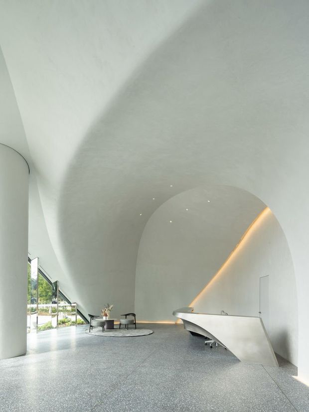 A dramatic minimalist interior with a soaring curved, barrel-vaulted white ceiling and sculptural arched wall forms. The space features a polished speckled grey floor, a sculptural angular reception desk, integrated warm linear lighting, small recessed ceiling lights, and a compact lounge seating area beside full-height glazing. The image appears to show a refined lobby or reception space with strong contemporary architectural detailing.