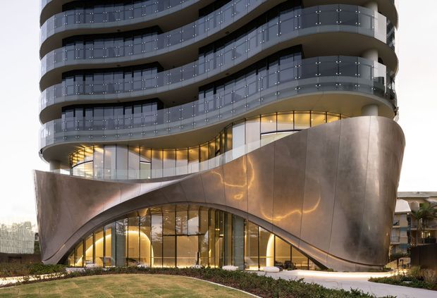A contemporary multi-storey apartment or mixed-use building with dramatically curved, continuous balconies, transparent glass balustrades, and a sculptural metallic podium facade. The lower level features sweeping arched openings and warm illuminated glazing, with landscaped planting in the foreground.