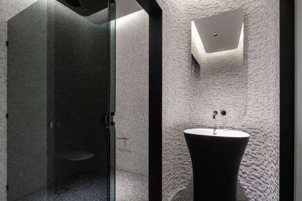 A compact, dramatic bathroom featuring a glass-enclosed shower on the left and a sculptural black pedestal basin on the right. Small grey mosaic tiles cover the walls and floor, with a mirrored recessed wall and concealed lighting creating a moody, high-contrast atmosphere.