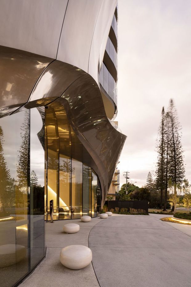 A dramatic contemporary building entrance viewed from the exterior, featuring sweeping sculptural metallic canopy forms, floor-to-ceiling glazed walls, a curved multi-storey facade, and a broad pale concrete forecourt with rounded stone seating. Warm interior lighting reflects through the glazing, while landscaped planting and tall narrow trees frame the approach.