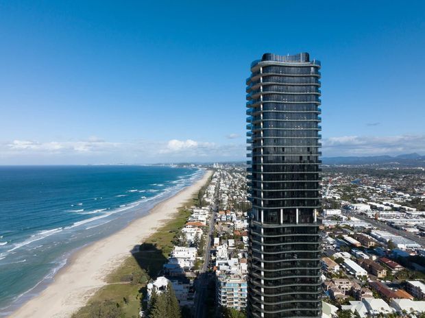 Aerial view of a tall, slender residential apartment tower with rounded corners, extensive dark glazing, and continuous horizontal balconies, positioned directly beside a long sandy beachfront. A dense coastal urban neighbourhood and distant mountain range extend behind it under a clear blue sky.