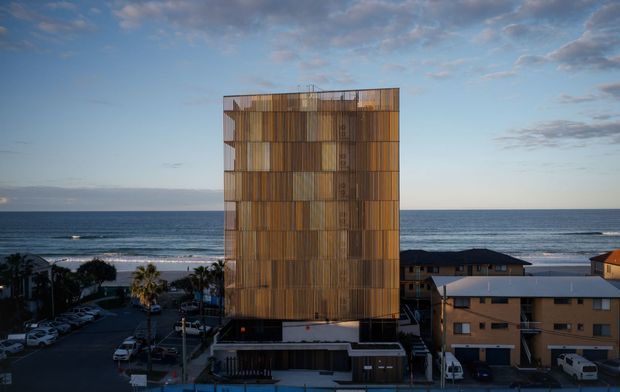 A tall, narrow multi-storey building viewed from across a coastal urban street, with a distinctive warm timber-toned vertical facade and rooftop railing. The ocean and horizon are directly behind the building, with low-rise neighbouring buildings and streetscape in the foreground.