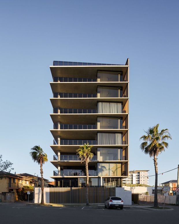 Street-level view of a contemporary multi-storey apartment building with stacked projecting balconies, extensive dark-framed glazing, warm timber-toned soffits and vertical screening. Palm trees, low-rise neighbouring buildings and a clear sky surround the urban residential facade.