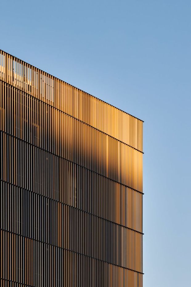 Close-up exterior view of a tall contemporary building facade, composed of closely spaced vertical timber-toned fins or slats arranged in layered horizontal sections. Warm sunlight highlights the upper portion, while darker shaded areas create strong geometric contrast against a clear blue sky.