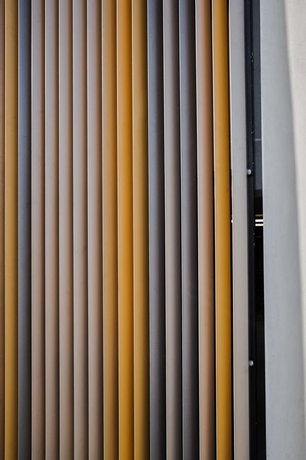 Close-up detail of a vertically ribbed architectural screen or facade composed of alternating narrow projecting strips in muted beige, grey, charcoal, and ochre-yellow tones. A black metal edge or frame is visible at the right beside a smooth pale wall. The image focuses on the repetitive textured facade treatment rather than a room or broader building context.