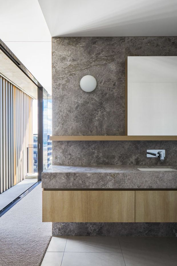 Contemporary bathroom vanity with a grey stone-clad feature wall, integrated stone benchtop and backsplash, timber floating cabinetry, round wall light, chrome wall-mounted tap, and large mirror. The bathroom opens to a bright glazed area with vertical screening and an urban outlook.