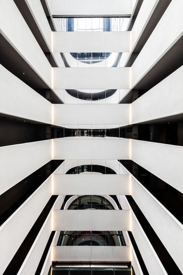 Vertically framed view looking through a multi-storey interior atrium, with repeated white balcony or corridor bands, dark recessed side walls, reflective glazing, and warm linear lighting. The symmetrical composition emphasizes the building’s substantial vertical void and contemporary commercial architecture.