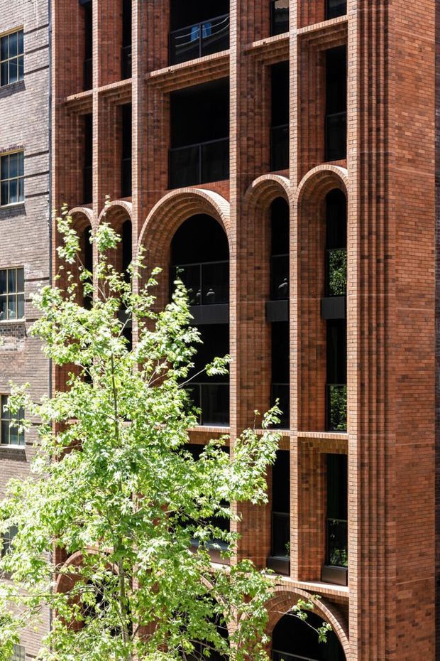 A close-up view of a multi-storey urban building facade clad in warm red-brown brick. The elevation features strong vertical brick piers, repeated tall recessed openings, dark-framed glazing, projecting balconies, and a series of prominent rounded brick arches. A leafy street tree partially obscures the lower facade.