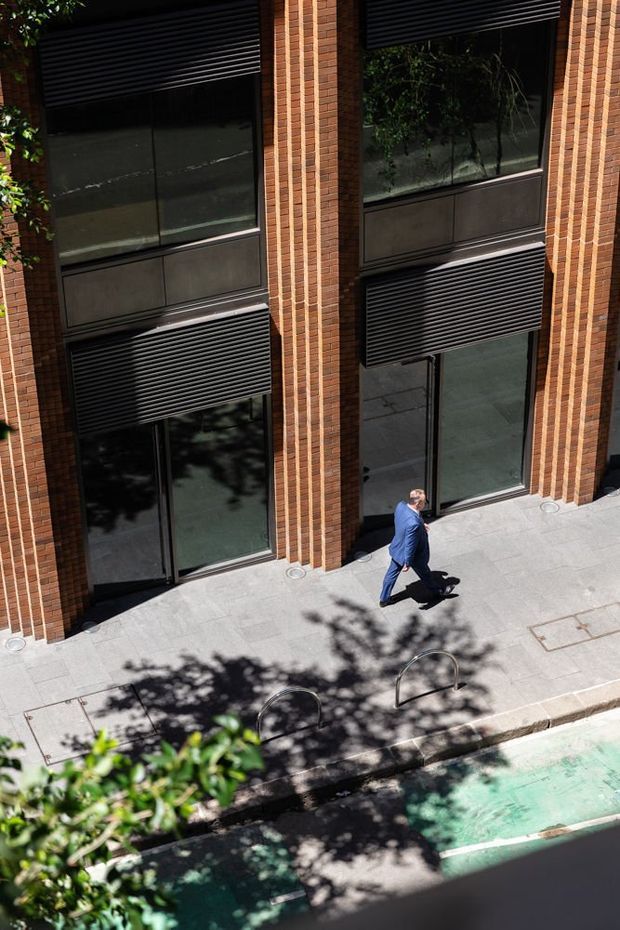 A street-level exterior view of a contemporary brick-clad multi-storey building, featuring tall dark-framed windows, external horizontal louvres or blinds, and a paved pedestrian forecourt with bicycle hoops. A person is walking along the frontage, while trees cast shadows across the pavement.