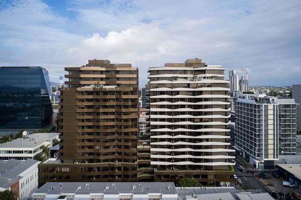 Aerial urban view of two prominent multi-storey apartment buildings with contrasting facades: one dark bronze/brown with regular horizontal balconies and the other light grey with distinctive undulating balcony edges. Surrounding commercial and residential high-rises, flat rooftops, and a partly cloudy sky are visible.