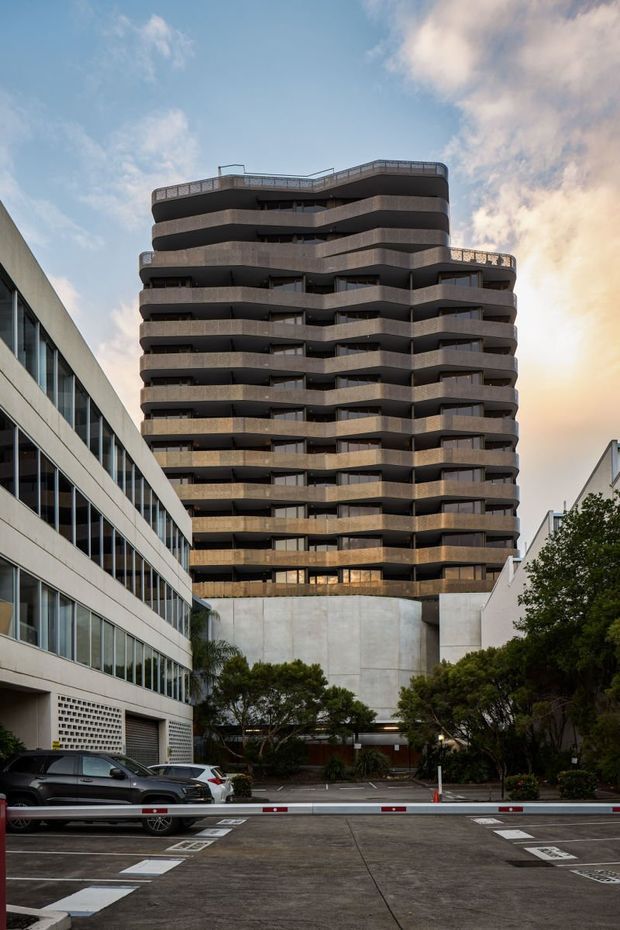 Exterior view of a tall contemporary multi-storey apartment building with rounded, undulating balconies and dark horizontal bands. The building rises above a concrete podium and landscaped driveway, with neighbouring commercial and residential structures visible on either side.