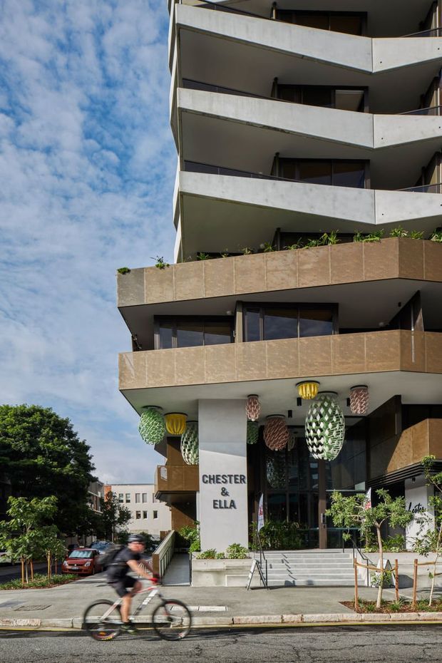 Street-level view of a contemporary multi-storey apartment or mixed-use building with projecting balconies, white concrete slab edges, bronze-toned balcony cladding, dark glazing, and a landscaped entrance. The ground-floor frontage is signed “CHESTER & ELLA” and features colourful hanging decorative lights. A cyclist and street trees are visible in the foreground.