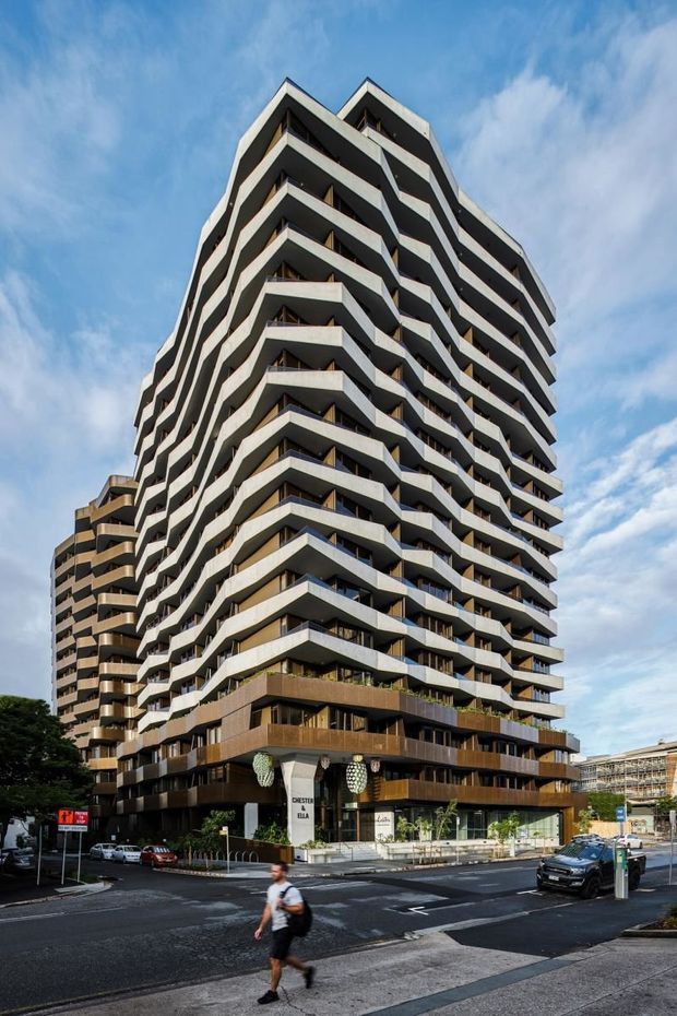 Street-level exterior photograph of a tall contemporary residential apartment building with strongly articulated stacked balconies, angular projecting slab edges, dark glazing, and a darker podium containing ground-floor commercial or lobby spaces. A second apartment tower and surrounding urban streetscape are visible in the background.