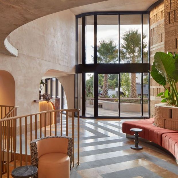 A refined, sunlit interior lobby or living space with a dramatic double-height glazed wall, black-framed floor-to-ceiling windows, curved textured plaster walls, patterned stone flooring, and warm timber and terracotta-toned seating. A sculptural staircase railing and large indoor plants contribute to the sophisticated contemporary atmosphere, with a landscaped courtyard visible beyond the glazing.