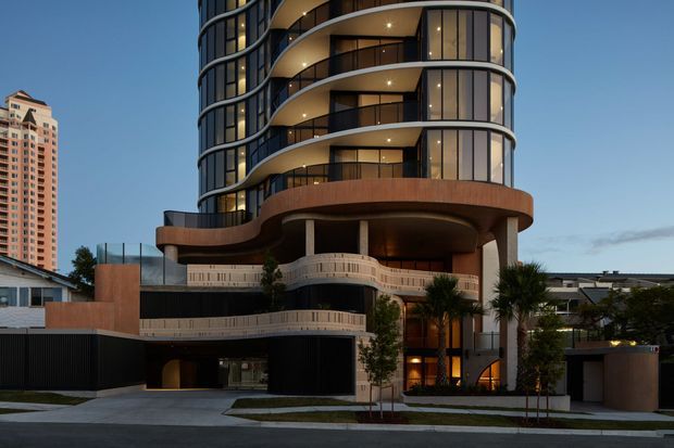 Contemporary multi-storey residential apartment building with a sculptural curved facade, rounded balconies, extensive dark-framed glazing, warm timber-toned cladding, and landscaped entry elements. The street-level view includes a recessed garage or vehicle entry and adjacent low-rise buildings.