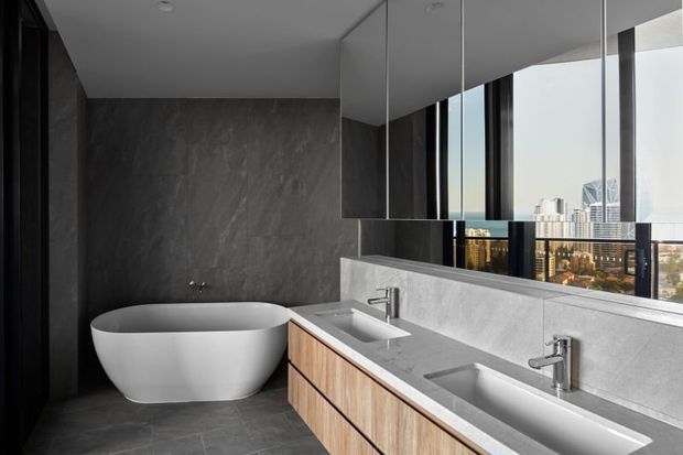 Contemporary bathroom with a freestanding white bathtub, double vanity, timber cabinetry, grey stone-look wall and floor finishes, and a large mirrored wall reflecting expansive city views through floor-to-ceiling glazing.