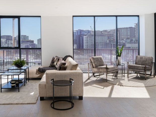 Bright contemporary living room with a beige sofa, two upholstered lounge chairs, glass-and-black metal coffee tables, a pale area rug, and a potted plant. Large black-framed windows provide expansive views of a dense urban apartment skyline and cast strong sunlight and shadows across the room.