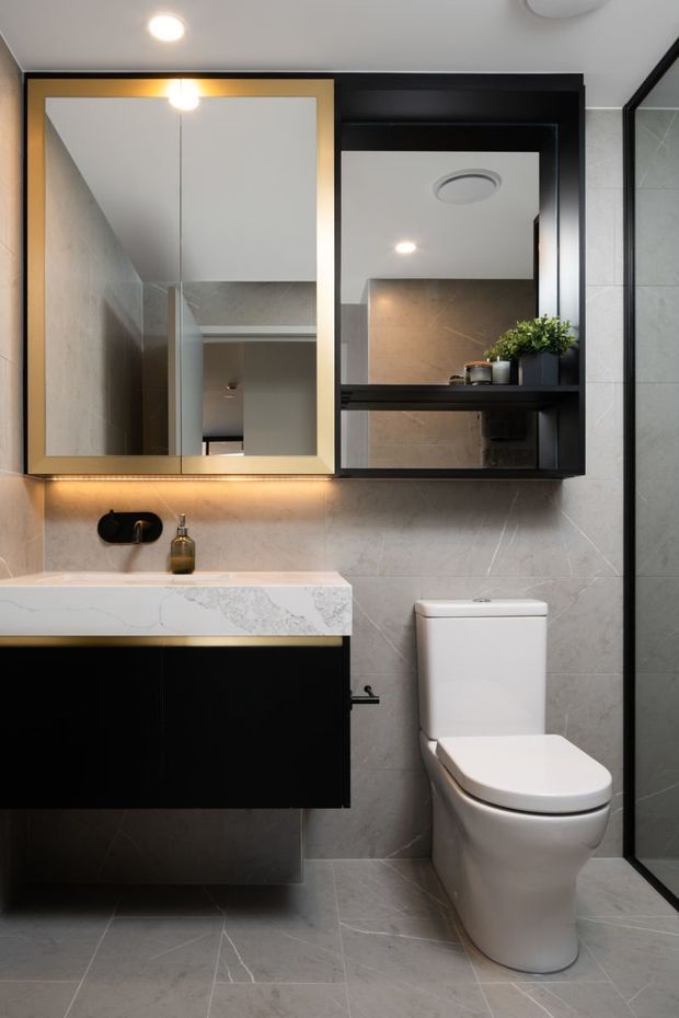 A compact contemporary bathroom featuring grey marble-look wall and floor tiles, a floating black vanity with a white veined stone benchtop, black-framed mirrored cabinetry with warm integrated lighting, a white toilet, and a black-framed glass shower enclosure. A small plant and toiletries add subtle styling.