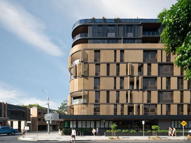 Street-level exterior view of a contemporary multi-storey apartment building with a rounded corner form, dark metal and glass upper levels, warm tan vertical facade panels, projecting balconies, and ground-floor commercial frontage. The building sits in an urban streetscape with pedestrians, street signage, and mature trees.