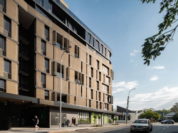 Street-level exterior view of a large contemporary multi-storey building with a curved corner facade, dark recessed window bays, warm timber-toned cladding, extensive glazing, and a ground-floor entrance marked “FABRIC”. The building fronts an urban street with pedestrians, street lighting, and nearby low-rise structures.