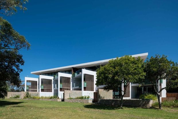 Exterior view of a contemporary low-rise residence with a broad asymmetrical sloping roof, white rendered walls, stone-clad lower sections, timber privacy screens, and extensive glazing. Mature trees and lawn frame the building under a clear blue sky.